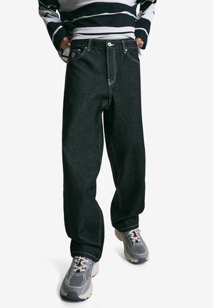 Persoon in ruime zwarte jeans met witte stiksels, grijs en witte sneakers, en een zwart-wit gestreepte lange mouwen shirt, staand met handen in de zakken.