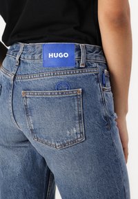 Laheljave denim kavbojke v svetlo modri barvi z rahlo zbledelim videzom, ki vključujejo modro HUGO logotipno nalepko na pasu in majhen okrogel logotip na žepu.