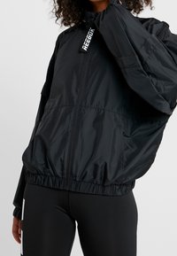 Chaqueta cortavientos negra con cierre de cremallera en el frente, puños y dobladillo elásticos, que presenta un collar alto y detalle de logotipo reflectante en el cuello.