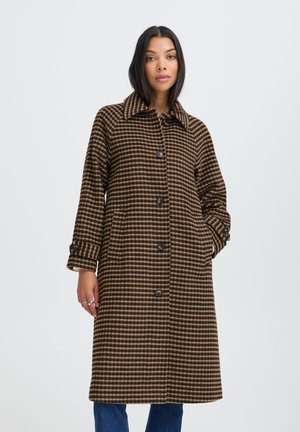 Femme portant un long manteau marron à carreaux boutonné avec col, debout avec une main dans la poche devant un fond clair uni.