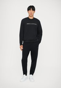 Schwarzer Sweatshirt mit dem Logo "ARMANI EXCHANGE", kombiniert mit schwarzen Hosen und weißen Sneakers. Einfaches Design, lockere Passform, glatte Stofftextur.