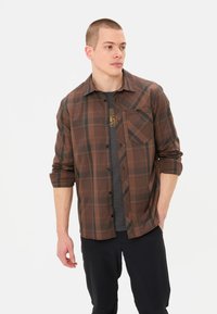 Bruine geruite shirt met een knoopsluiting aan de voorkant, één borstzak en lange mouwen opgerold bij de manchetten. Draag het over een grijs T-shirt.