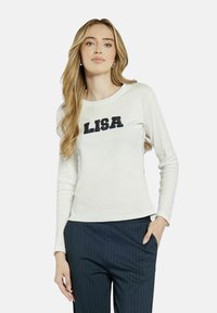 Długi rękaw, kremowa bluzka z okrągłym dekoltem, na piersi z napisem "LISA" w czarnym kolorze. Gładka tekstura.