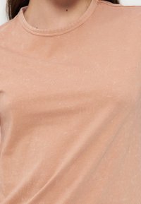 Κοντινό πλάνο ενός ατόμου που φοράει ξεθωριασμένο ροδακινί κοντομάνικο μπλουζάκι με στρογγυλή λαιμόκοψη και διακριτή υφή.