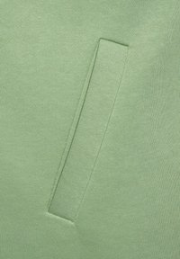 Tessuto a maglia verde con una finitura testurizzata presenta un dettaglio di tasca rettangolare, integrato nella superficie. L'aspetto complessivo è liscio e senza interruzioni.