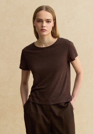 Jeune femme aux cheveux blonds et raides porte un t-shirt marron foncé à manches courtes et un pantalon assorti, debout les mains dans les poches.