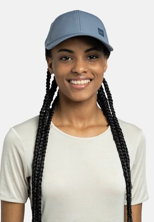 Joven con cabello largo trenzado, que lleva una camiseta gris claro de manga corta y una gorra de béisbol azul grisáceo, sonriendo frente a un fondo liso.