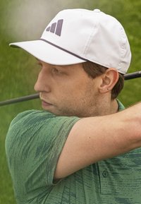 Casquette blanche avec un accent sombre, logo à trois bandes, associée à un polo vert texturé. Le polo a des manches courtes et un col.