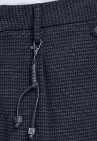 Mörkt mönstrat tyg med texturerad yta, utrustat med dragkedja och justerbara dragsnören, kompletterat med metalldetaljer.