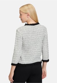 Weißer und schwarzer Tweed-Jacke mit strukturiertem Muster, schwarzen Fransen-Akzenten am Kragen und an den Bündchen, 3/4-Ärmeln und taillierter Passform.