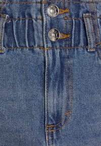 Blå denimtyg med en texturerad yta, som har två metallknappar, kontrasterande gul sömnad och en rynkad elastisk midja.