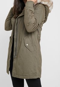 Olivegrüner Parka mit Reißverschluss und Druckknopfverschluss, ausgestattet mit einer Kapuze aus Kunstpelz und zwei Fronttaschen. Texturiertes Material.
