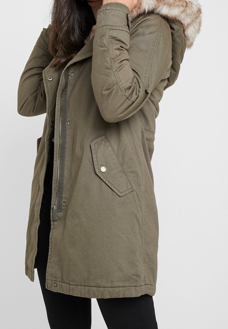 Olivegrüner Parka mit Reißverschluss und Druckknopfverschluss, ausgestattet mit einer Kapuze aus Kunstpelz und zwei Fronttaschen. Texturiertes Material.