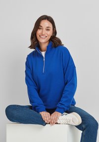 Royal blue sweatshirt with a high collar and quarter-zip, featuring a relaxed fit and ribbed cuffs, paired with blue jeans and white shoes.