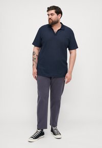 Navy striped polo shirt with short sleeves and a v-neck, paired with gray pants and black high-top sneakers. Features tattoos on the arm.