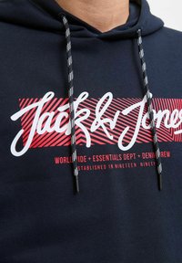 Granatowy hoodie z graficznym nadrukiem w czerwone paski, na którym znajduje się napis "Jack & Jones" w białym, kursywnym foncie. Akcenty w czerni i bieli przy sznurku. Materiał w mieszance bawełny.