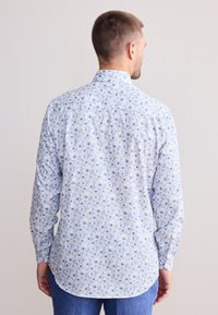 Muž v dlhom bielom tričku s modrým kvetinovým vzorom, zobrazený zozadu, v modrých nohaviciach na jednoduchom svetlom pozadí.