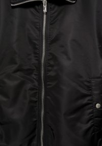 Veste noire avec une fermeture éclair centrale en métal argenté, une tirette de zip au design gravé, et un rabat de poche avec un bouton pression métallique.
