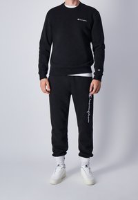 Sudadera negra con logo en el pecho, combinada con pantalones de chándal a juego que presentan el logo en el lateral. Lleva puestas zapatillas blancas y calcetines texturizados.