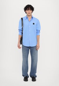 Himmelblaues Button-up-Hemd mit langen Ärmeln und Brusttasche, kombiniert mit hellblauen Jeans und schwarzen Leder-Schuhen. Schwarze Tasche über der Schulter.