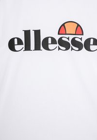 Vitt T-shirt med en tydlig svart "ellesse"-logotyp och en färgglad halvcirkel design i orange och röd ovanför texten. Slät tygkvalitet.