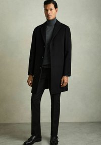 Sort uldfrakk med slagkrave, enkelt lukning, båret over en mørk turtleneck. Slim-fit sorte bukser og sko fuldender looket.