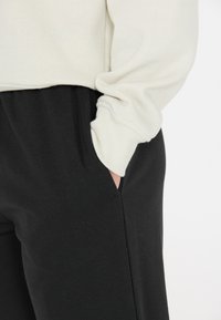 Sweatshirt i mjuk creamfärgad tyg med ribbade ärmslut, bärs tillsammans med svarta joggingbyxor med sidfickor och en slät yta.