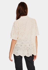 Beige kortärmad topp med volangkrage, som har utskurna blommönster och vågig nederkant. Peplum-siluett med texturerad tyg.