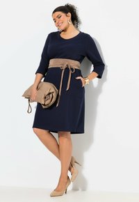 Marineblaues Kleid mit Dreiviertelärmeln, einem beige Stoffgürtel und vorderen Taschen. Kombiniert mit beige hohen Schuhen und einer gefalteten Clutch.