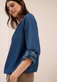 Femme portant une chemise bleue à manches longues aux manches retroussées et un pantalon marron, se tenant devant un fond beige uni.