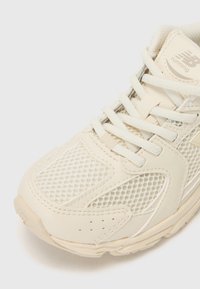 Lys beige løpesko med mesh-overdel, forsterket tå og polstret såle. Har lisser og logo på tungen for merkevarebygging.