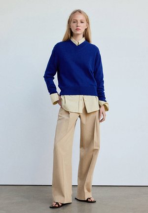 Jeune femme debout portant un pull bleu par-dessus une chemise beige avec un pantalon beige large et des sandales noires à bouts ouverts sur un fond uni.