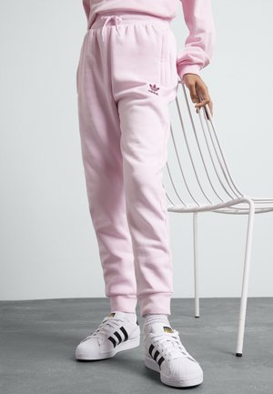 Osoba nosząca jasnoróżowe spodnie dresowe Adidas i białe buty Adidas stoi obok białego metalowego krzesła na szarej podłodze.