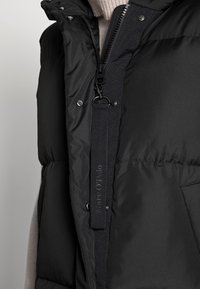 Gilet imbottito nero con colletto alto, dotato di chiusura zip, bottoni a pressione e etichetta di Marc O'Polo. Tessuto testurizzato, dettaglio tasca.
