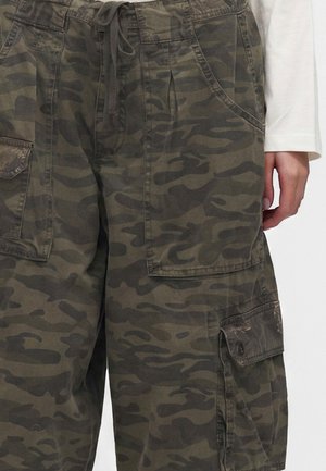 Close-up af person iført mørkegrønne og brune camouflerede cargobukser med store lommer og snøre i taljen, samt en hvid langærmet top.