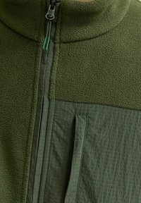 Chaqueta de forro polar verde con un cuello alto, que presenta un tejido texturizado en la parte superior y un panel liso con un patrón de cuadrícula y un bolsillo con cremallera.