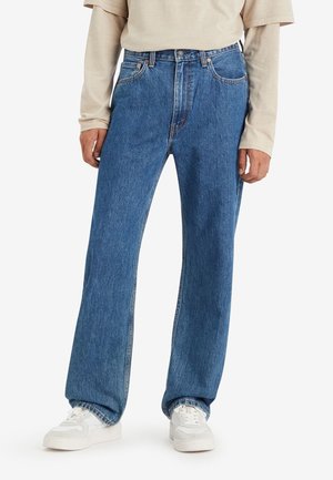 Personne portant un jean bleu ample et des baskets blanches, debout les mains relâchées le long du corps sur un fond blanc uni.