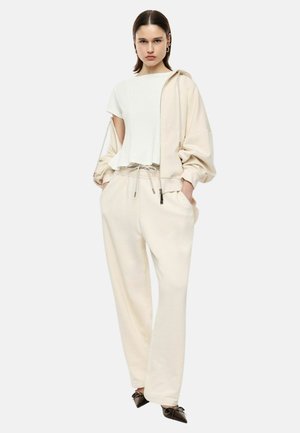 Donna che indossa una felpa color crema con cappuccio e pantaloni larghi, con top bianco e scarpe nere appuntite, in piedi con le mani in tasca.
