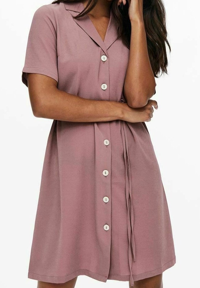 Femme portant une robe rose poudré à manches courtes, boutonnée, avec un col et une ceinture en tissu à la taille, debout avec un bras croisé.
