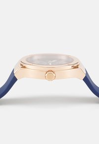 Reloj de pulsera con caja de oro rosa, esfera oscura, correa de silicona azul y una corona prominente. Presenta múltiples subesferas y un acabado luminoso.