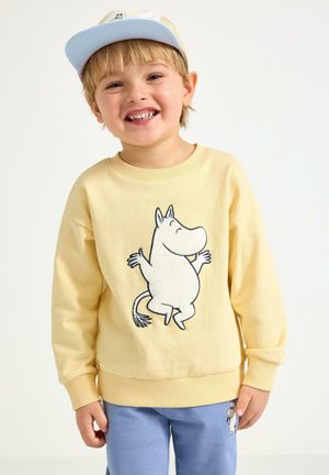 Gul bomullströja med Muminmotiv på framsidan. Matchas med blåa mjukisbyxor och en ljusblå keps. Mjuk textur.