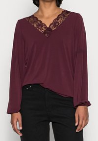 Burgundowa bluzka z długim rękawem, dekoltem w kształcie litery V i koronkowym wykończeniem przy dekolcie, o luźnym kroju i miękkiej fakturze tkaniny.