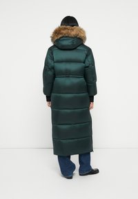 Abrigo inflable largo de color verde oscuro con capucha forrada de piel, que presenta un diseño acolchado y puños acanalados, que llega hasta la mitad de la pantorrilla.