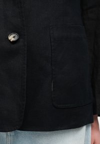 Μαύρο λινό σακάκι με ένα κουμπί και ένα ορθογώνιο εμπρόσθιο τσεπάκι, φορεμένο πάνω από ανοιχτόχρωμα τζιν παντελόνια.