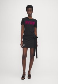 Svart t-skjorte med fuksia "Versace Jeans Couture"-logo, sammen med et svart miniskjørt med knyttet liv og hvit logo-detalj.