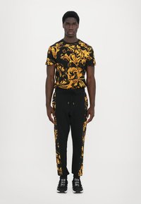 Black t-shirt with gold baroque pattern, paired with black joggers featuring gold accents and white side stripes. Shoes are black with white soles.