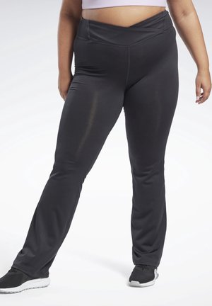 Personne portant un pantalon noir taille haute à jambes larges et des baskets noires, debout avec une jambe légèrement en avant sur un fond blanc.