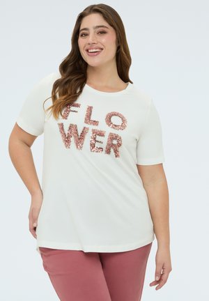 Mujer sonriente con largo cabello castaño que lleva una camisa blanca de manga corta con el texto "FLOWER" en lentejuelas rosas y pantalones rosas, posando contra un fondo liso.