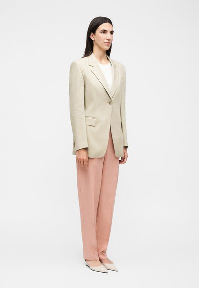 Γυναίκα που φοράει μπεζ σακάκι, λευκό τοπ, φαρδιά ροζ παντελόνια και μυτερές μπεζ παπούτσια σε απλό φωτεινό φόντο.