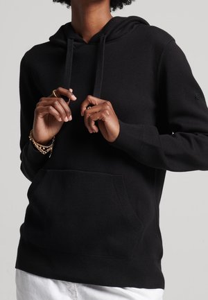 Person wearing a black hoodie with front pocket and drawstrings, adjusting the strings, accessorized with gold bracelets and rings.